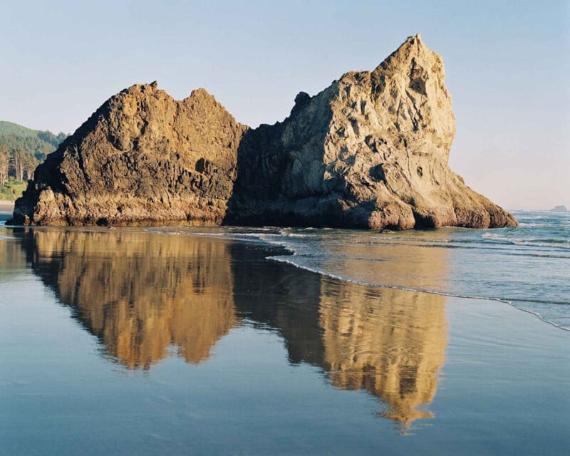 Enormes estructuras rocosas se elevan desde el borde del océano, claramente reflejadas en la arena húmeda durante la marea baja bajo un cielo despejado. Al fondo, a la izquierda, se puede ver la vegetación costera.