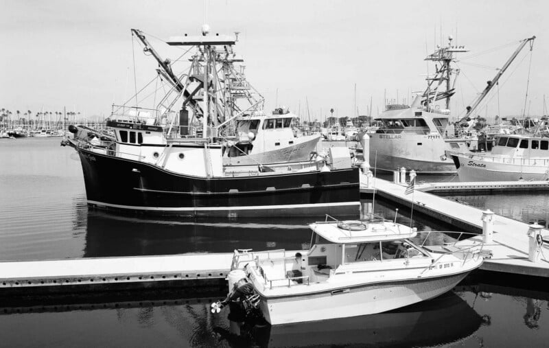 Hay varios barcos pesqueros atracados en el muelle, con un barco más pequeño en primer plano y barcos pesqueros comerciales más grandes detrás. El agua tranquila refleja los barcos y al fondo se puede ver la lejana costa.