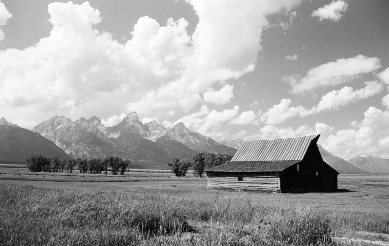 Una fotografía en blanco y negro de un granero rústico de madera en una pradera, con una hilera de árboles y montañas altas y escarpadas al fondo, y un cielo lleno de nubes dispersas.