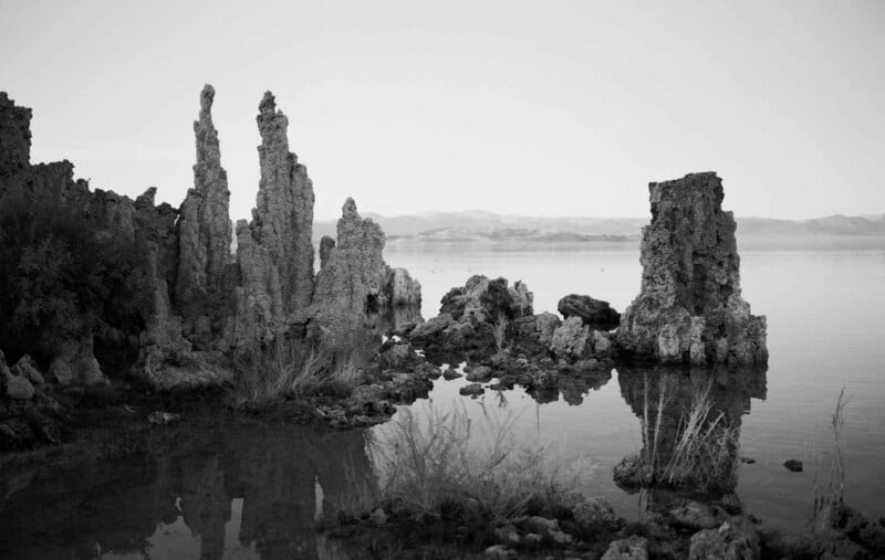 La imagen en blanco y negro muestra formaciones rocosas altas y escarpadas que se elevan desde el borde de un lago en calma, con montañas distantes y cielos despejados al fondo. Entre las rocas crece una escasa vegetación.