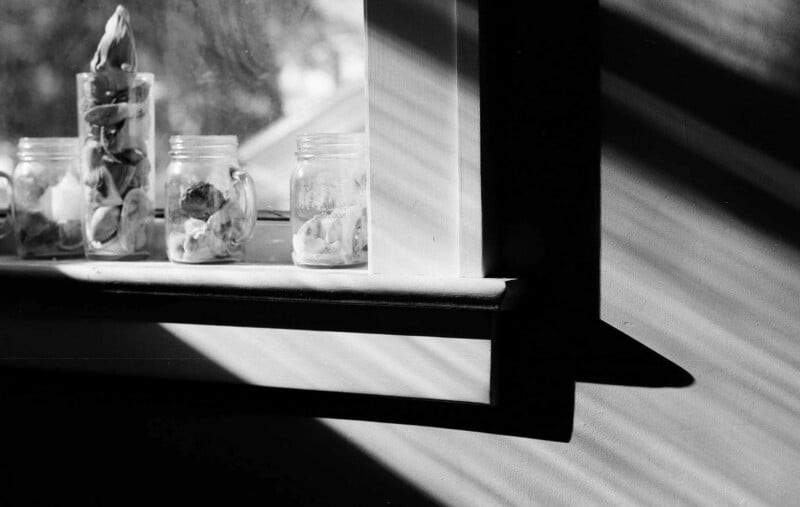 Frascos de vidrio llenos de conchas marinas se encuentran en el alféizar de la ventana, y la luz del sol brilla a través de las ventanas, proyectando sombras dramáticas y patrones de luz en las paredes. La escena es en blanco y negro.