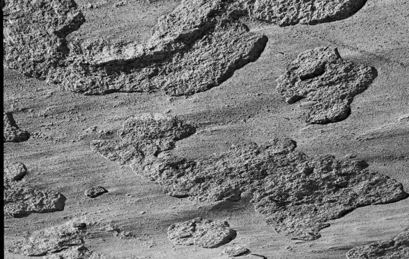 Bloques de hormigón o piedra rugosos y de textura desigual cubren una superficie plana y arenosa, creando un patrón abstracto de formas irregulares y sombras en la imagen en blanco y negro.