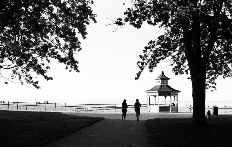 Dos personas caminaban por el sendero que conducía al pabellón de la playa, que estaba rodeado de grandes árboles. La escena es en blanco y negro, con un cielo brillante y figuras lejanas cerca de la orilla del agua.