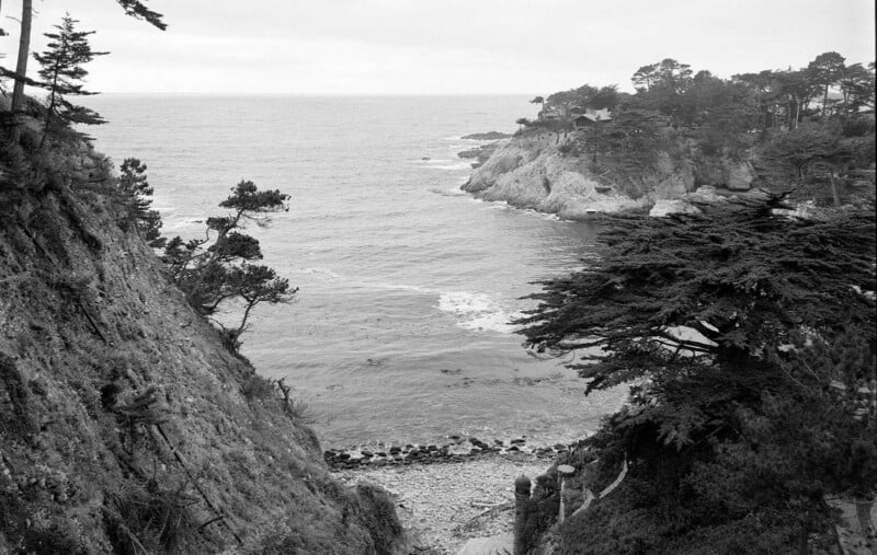 Una fotografía en blanco y negro que muestra una costa rocosa, acantilados, densos árboles y olas rompiendo suavemente en una playa de guijarros. Bajo un cielo nublado, el océano se extiende hasta el horizonte.