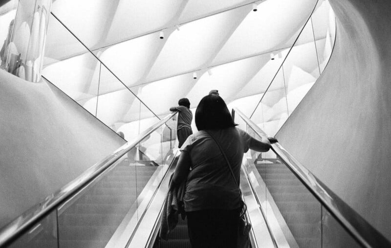 Un hombre sube por una escalera mecánica en un edificio moderno con mucha luz y un techo geométrico. Otro hombre estaba delante, apoyado contra la barandilla de cristal. La imagen está en blanco y negro.