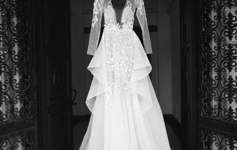 Un vestido de novia blanco con mangas largas de encaje y detalle de falda escalonada colgaba en la entrada, rodeado por una ornamentada puerta de hierro forjado.