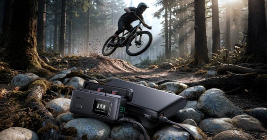 A mountain biker jumps over rocks on a forest trail, with a portable charging device and camera batteries in focus on the rocky ground in the foreground. Sunlight filters through tall trees.