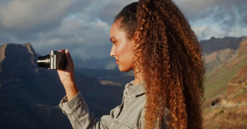 Una mujer de pelo largo y rizado toma una fotografía al aire libre con una cámara, con montañas y un cielo parcialmente nublado al fondo.