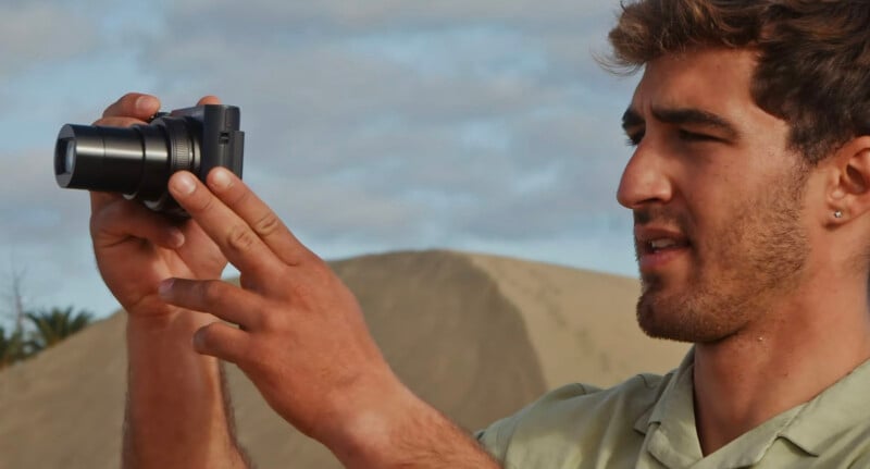 Un hombre sostiene una cámara digital liviana al aire libre y se concentra en tomar fotografías. Detrás de él, hay una montaña de arena y un cielo despejado.