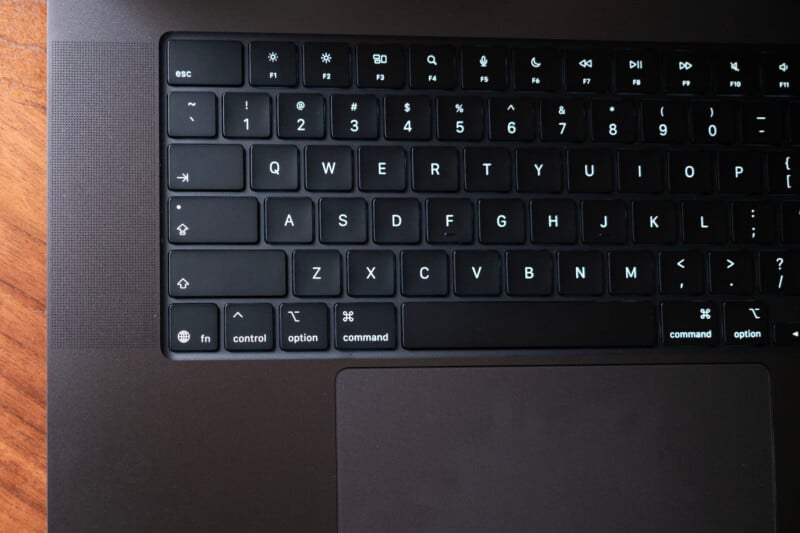 A close-up of the left side of a black laptop keyboard, showing illuminated keys including control, option, command, and function, with part of the trackpad visible below. The laptop rests on a wooden surface.