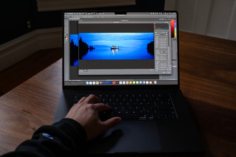 A person’s hand on a laptop keyboard with a photo editing program open, displaying a vivid blue landscape image of a lake with trees and a boat, on a wooden table indoors.