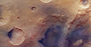 A high-resolution image of the Martian surface shows several impact craters of varying sizes on a dusty, reddish-brown landscape with some shadowed and rough textured areas.