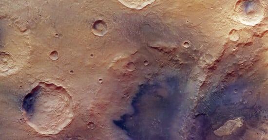 A high-resolution image of the Martian surface shows several impact craters of varying sizes on a dusty, reddish-brown landscape with some shadowed and rough textured areas.