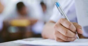 Close-up of a person’s hand holding a pen and writing on paper, with a blurred background of other people, suggesting a classroom or exam setting.