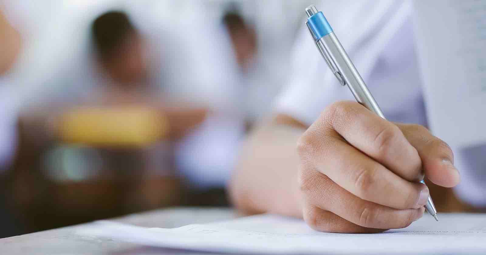 Close-up of a person’s hand holding a pen and writing on paper, with a blurred background of other people, suggesting a classroom or exam setting.