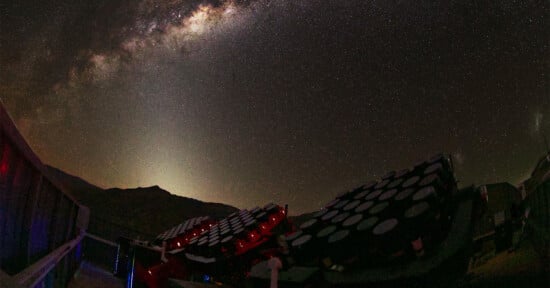 Telescope arrays with circular lenses are pointed toward a star-filled night sky, with the bright band of the Milky Way visible above distant mountain silhouettes.