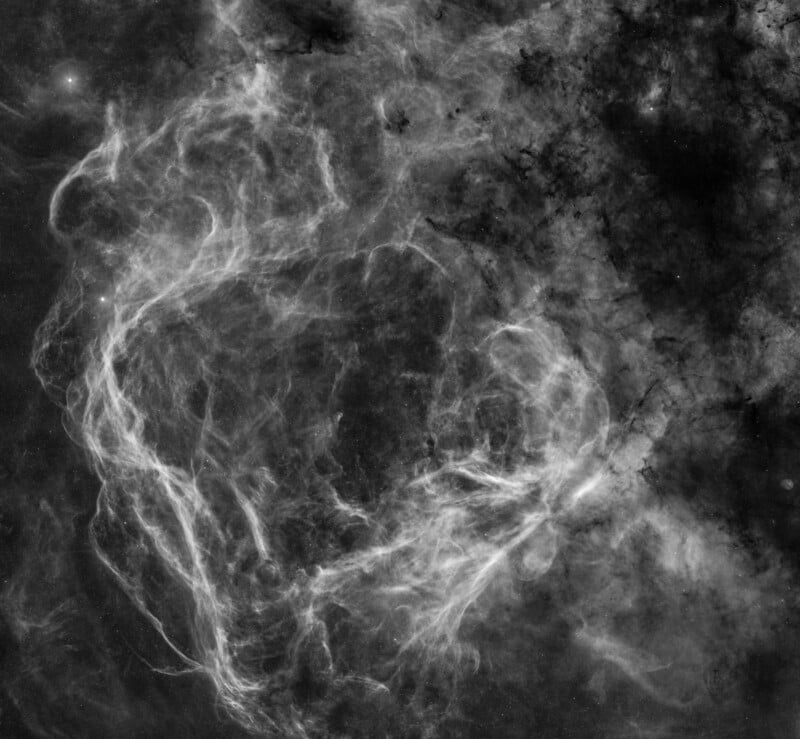 Black-and-white image of wispy, cloud-like filaments in space, resembling tangled smoke or nebula structures, set against a dark background with scattered bright spots.
