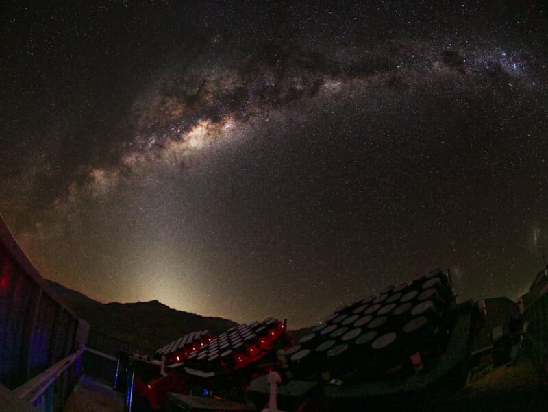Vista nocturna de gran angular de un telescopio bajo un cielo despejado, con la Vía Láctea arqueándose en lo alto y estrellas densamente esparcidas en el cielo oscuro sobre un paisaje montañoso.