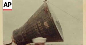 A close-up of a NASA space capsule being hoisted by a cable, with a sailor in a white hat partially visible in the foreground. The AP logo appears in the top left corner.