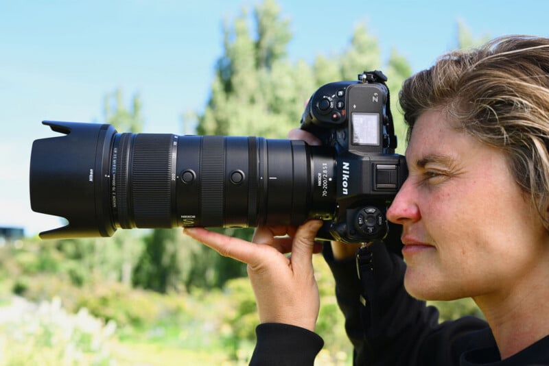 A person holds a Nikon camera with a large telephoto lens, aiming it at something in the distance outdoors, with green trees and a blue sky in the background.