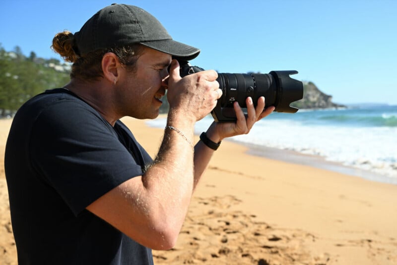 A person wearing a black shirt and cap is taking photos with a camera on a sunny beach, with waves and rocky hills visible in the background.