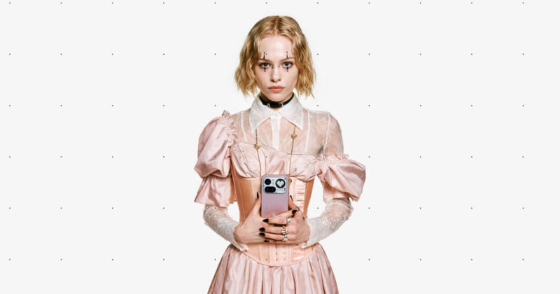 A woman with short blonde hair, wearing a pale pink, vintage-style dress with puffed sleeves and a lace collar, stands against a plain background, holding a smartphone with both hands in front of her.