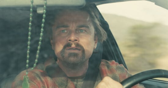 A man with a beard and mustache sits behind the wheel of a car, looking ahead with a concerned expression. Green beads hang from the rearview mirror, and sunlight illuminates the scene through the windshield.