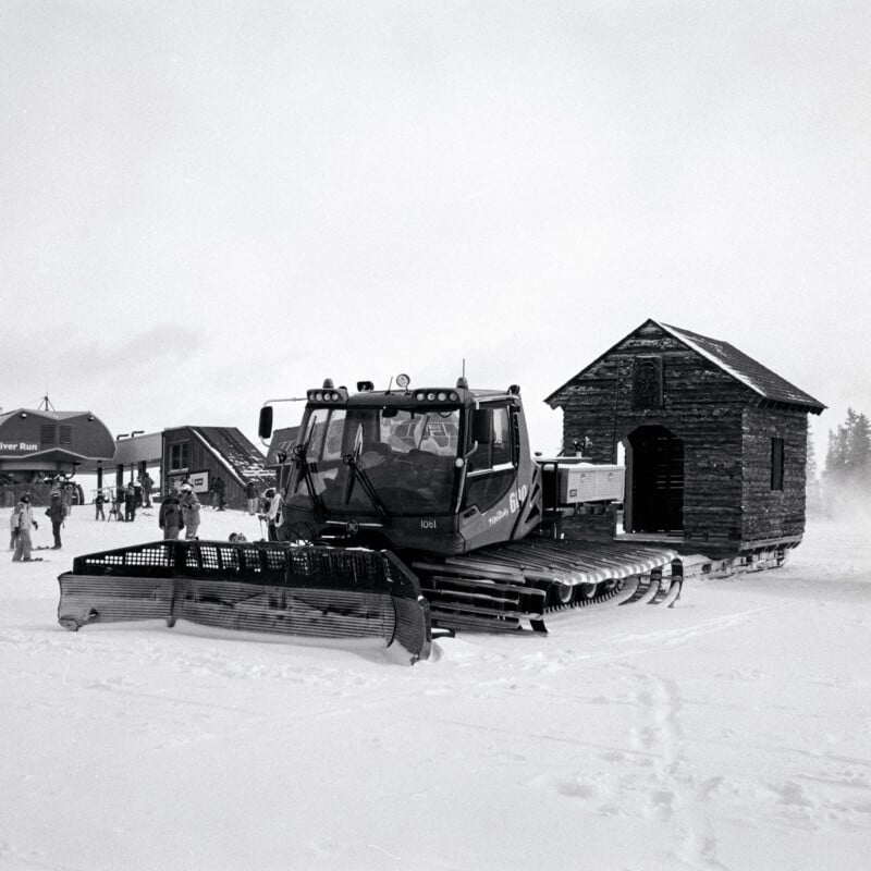 A snowcat vehicle pulls a small wooden cabin on a snowy landscape, while people stand nearby at a ski area with buildings in the background.