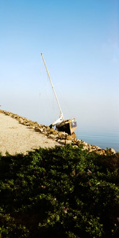 A sailboat is tilted on its side among rocks near a sloped shoreline under a clear blue sky. Green shrubbery is visible in the foreground.