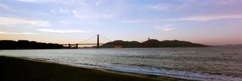 A wide view of the Golden Gate Bridge at sunset, seen from a sandy beach with gentle waves, with hills and pastel sky in the background.