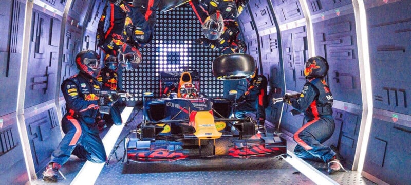 A Formula 1 pit crew in Red Bull uniforms works on a race car in a futuristic, tunnel-like garage, with crew members positioned at various points around the vehicle.