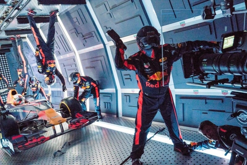 Race car pit crew working on a Formula 1 car inside a futuristic, enclosed room; some crew members appear to be standing or working upside down, and a cameraman films the scene.