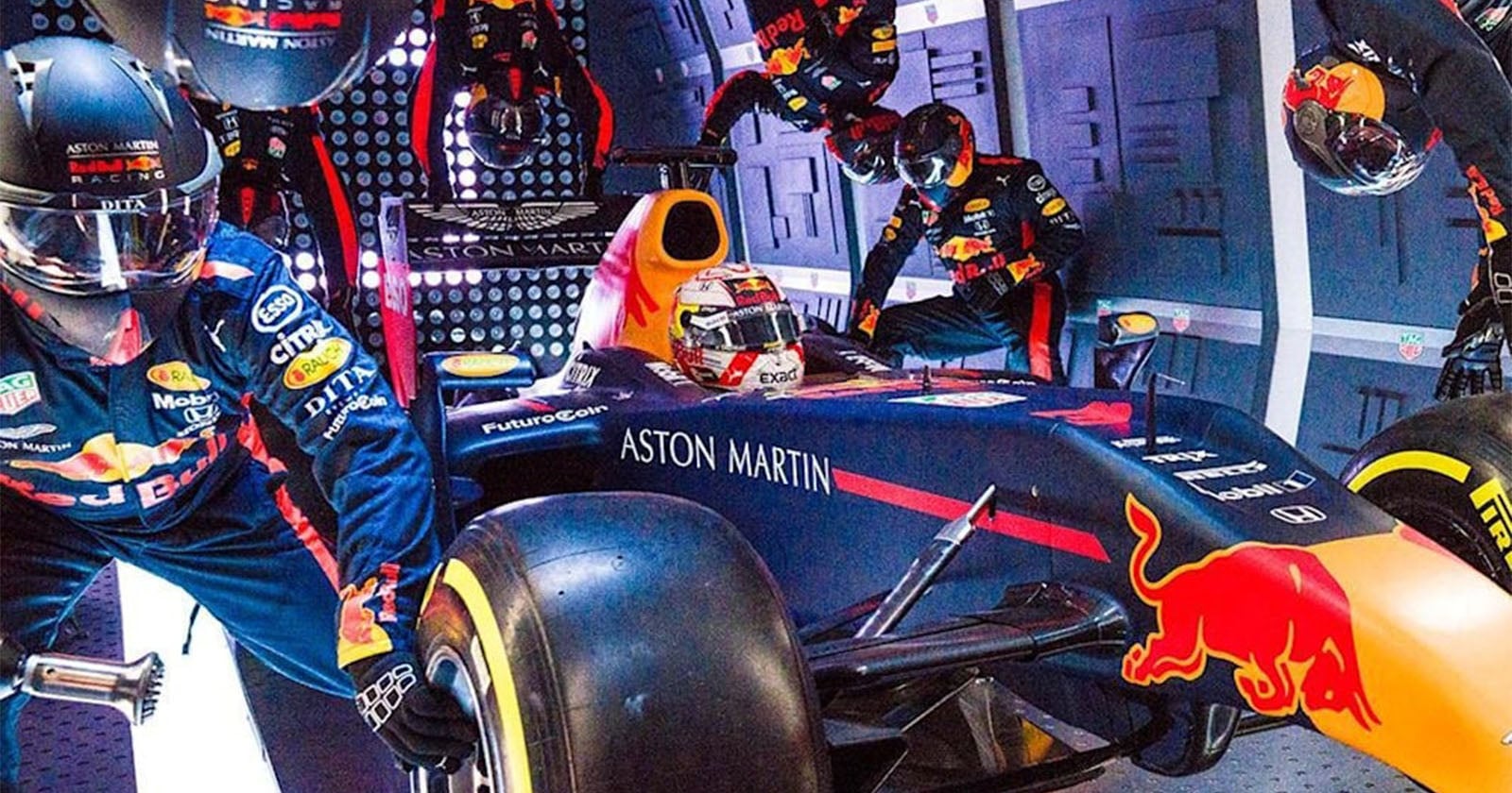 A Formula 1 Red Bull racing car stopped in the pit lane, surrounded by a pit crew in helmets and racing suits, as they perform a quick tire change and maintenance.