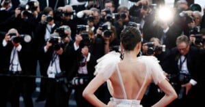A woman in a glamorous dress poses with her back to the camera on a red carpet, facing a crowd of photographers taking pictures with bright flashes.