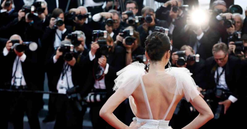A woman in a glamorous dress poses with her back to the camera on a red carpet, facing a crowd of photographers taking pictures with bright flashes.