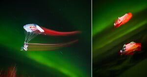 A parachutist with a Red Bull parachute glides through a night sky illuminated by green auroras, with red light trails behind them. On the right, two parachutists in red suits are seen against the glowing sky.