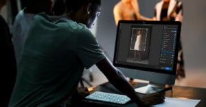 A person uses a desktop computer to edit a photo of a woman in a white dress; other people and studio lighting equipment are visible in the blurred background.