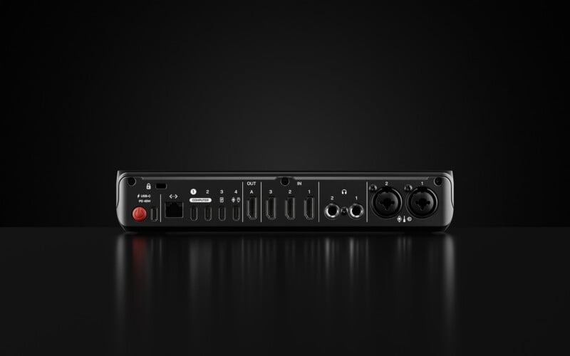 A black audio interface with multiple ports and input/output connections on the back, including HDMI, USB, headphone jacks, and audio inputs, displayed against a black background.
