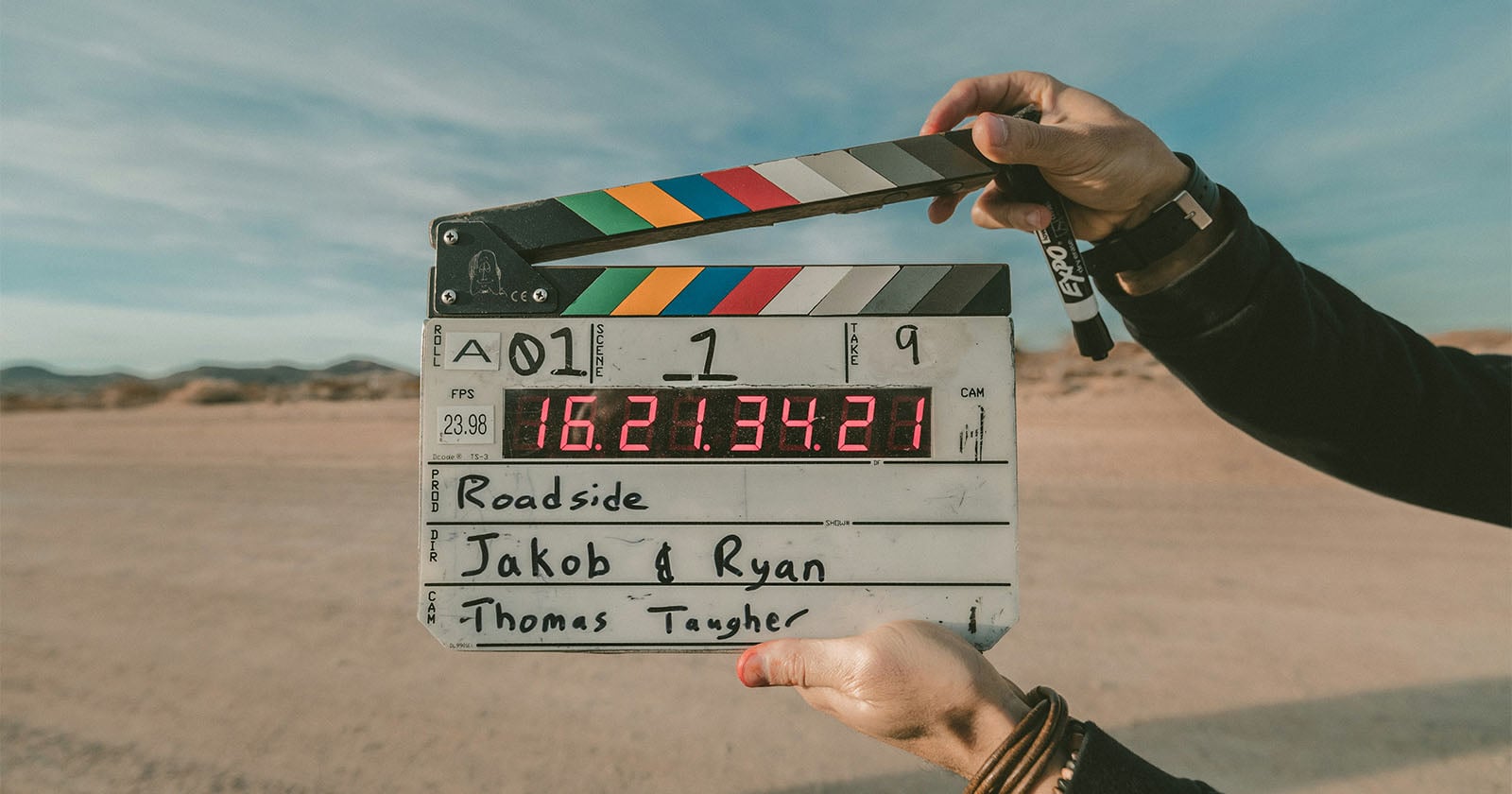 A person holds a film clapperboard with colorful stripes and a digital timecode display, standing outdoors on a sandy landscape with mountains and a blue sky in the background.