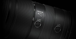 Close-up of a black Nikkor S camera lens, showing detailed texture, focal length markings (135, 200), and two customizable L-Fn buttons on the lens barrel against a dark background.