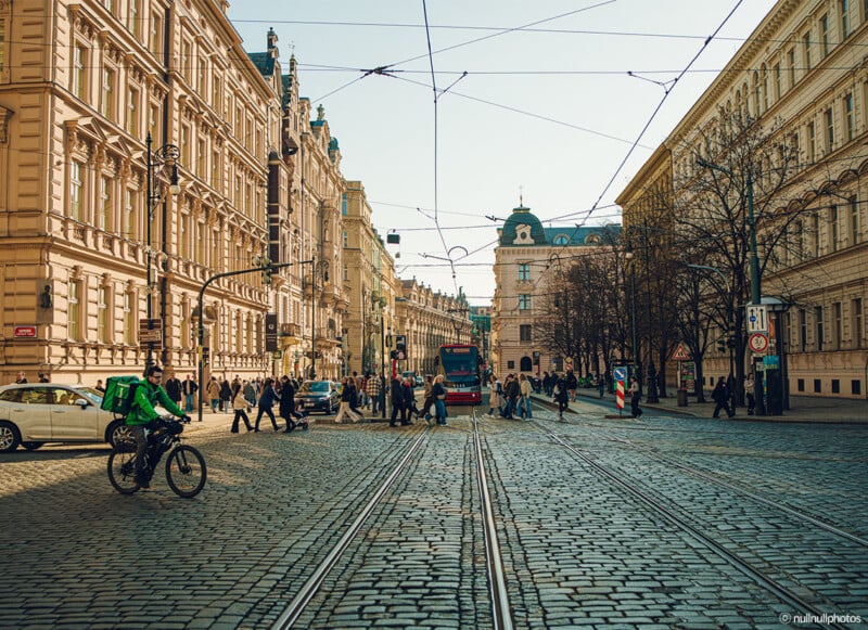 A busy European city street with cobblestone paving, intersecting tram tracks, people walking, a cyclist, and a red tram. Historic buildings line both sides under a clear sky with visible overhead tram wires.
