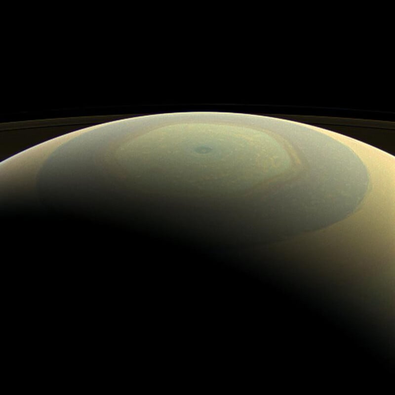 A view of Saturn's north pole shows a large, distinct, hexagonal weather pattern surrounded by swirling clouds, with part of the planet in shadow against the dark background of space.