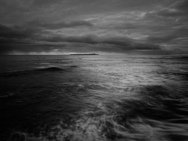 A black and white photo of a choppy sea under dramatic, cloudy skies, with an island and lighthouse visible on the horizon in the distance.