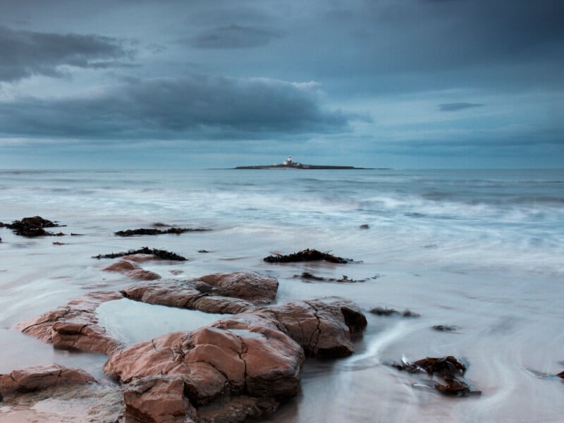 Rocky shoreline with seaweed in the foreground, gentle waves, and an island with a lighthouse in the distance under a cloudy, blue-grey sky.