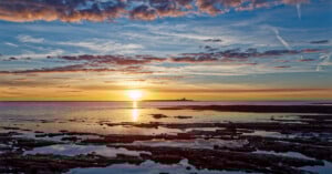 A vibrant sunset over a calm sea with rocky tide pools in the foreground and a distant silhouette of an island or lighthouse on the horizon, beneath a sky filled with colorful clouds.