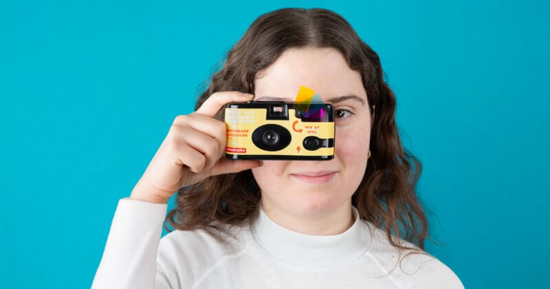 A woman with wavy brown hair and a white turtleneck holds a yellow disposable camera up to her face, smiling, against a bright blue background.