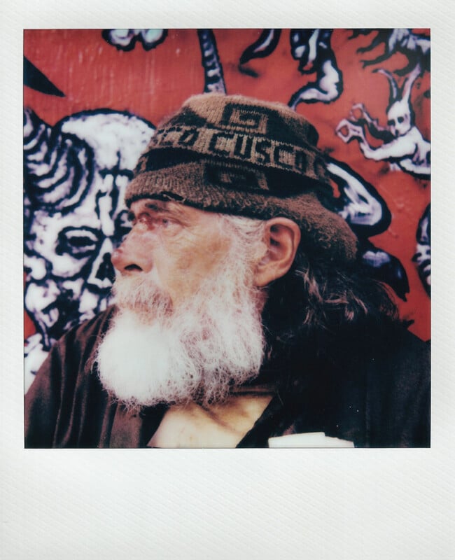 Un anciano con una espesa barba blanca y cabello largo, que lleva un gorro de punto estampado, mira junto a un vivo mural rojo de calaveras y criaturas extrañas.