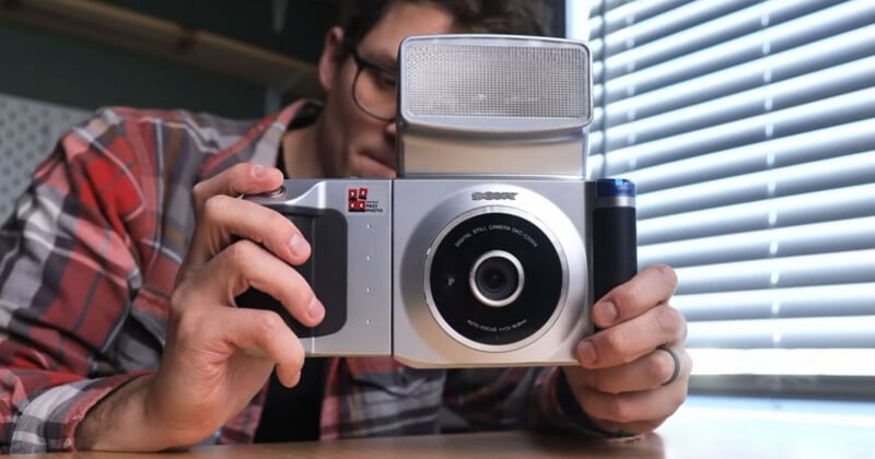 A person wearing glasses and a plaid shirt holds a large, retro-style Sony digital camera with a prominent lens and built-in flash, near a window with blinds.
