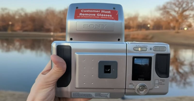 A hand holds a vintage Sony digital camera outdoors by a lake; a red sticker on the camera reads, "Customer Must Remove Glasses (per US Department of State – effective 11/1/16)." Trees and grass are in the background.