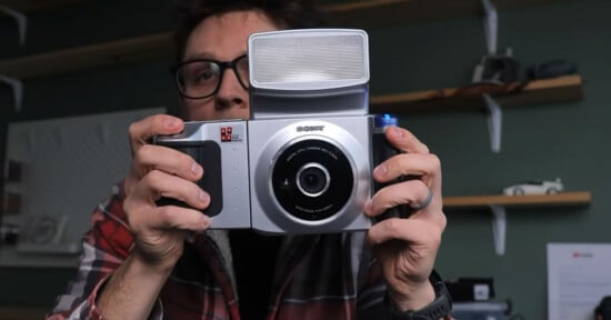 A person in glasses and a plaid shirt holds a large, vintage-style digital camera with a prominent flash, facing toward the camera in a room with shelves and various objects in the background.